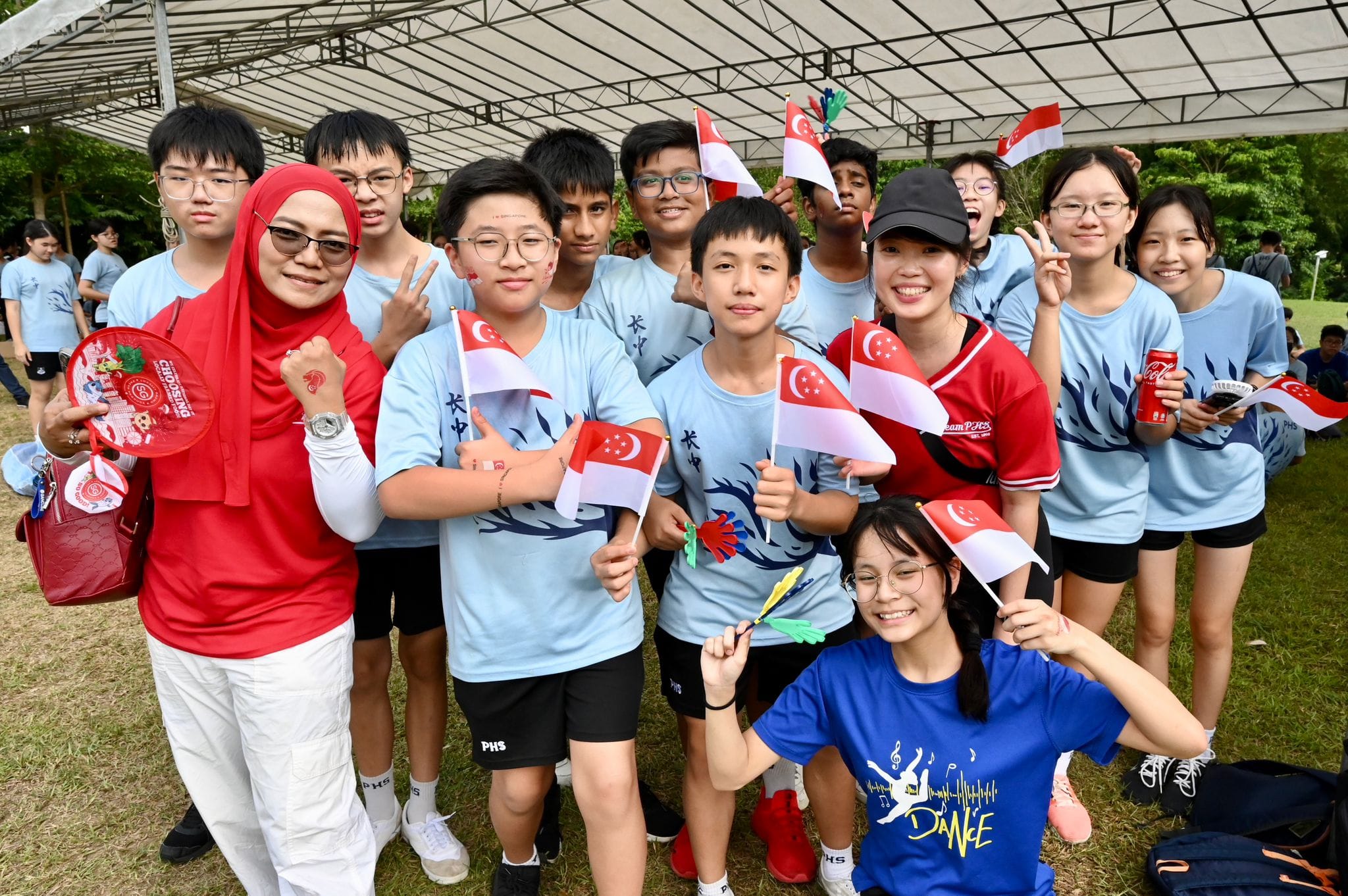 A group of smiling students in light blue shirts wave Singapore flags at an outdoor event under a tent.